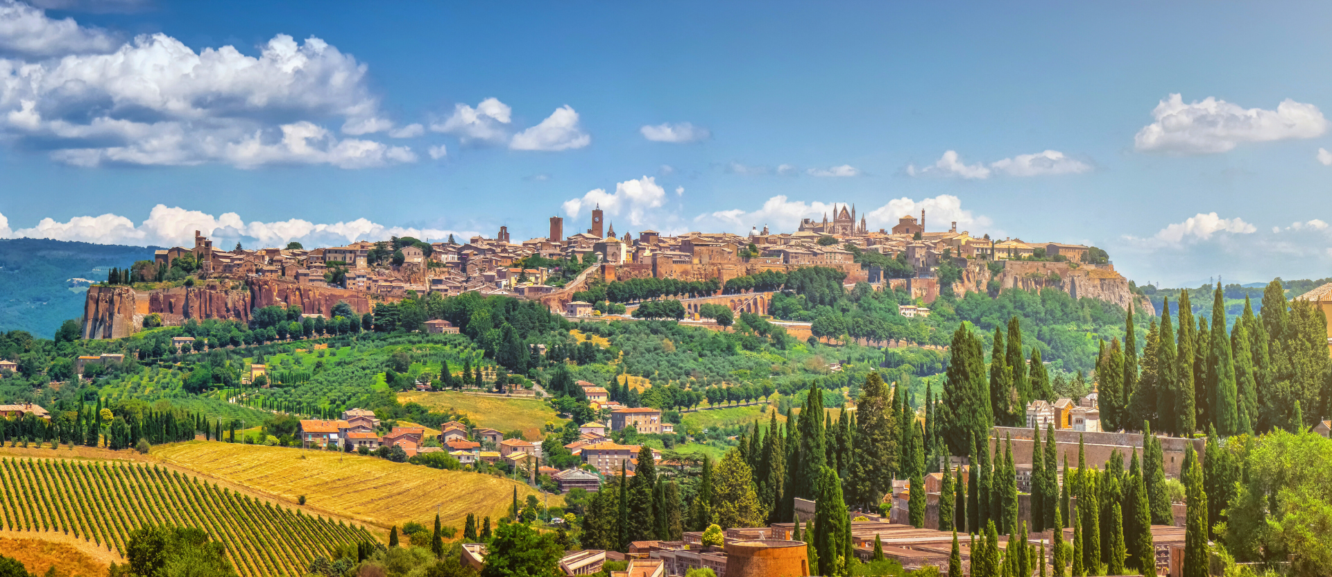 Italian hilltop town surrounded by trees and countryside beneath a blue sky with white clouds