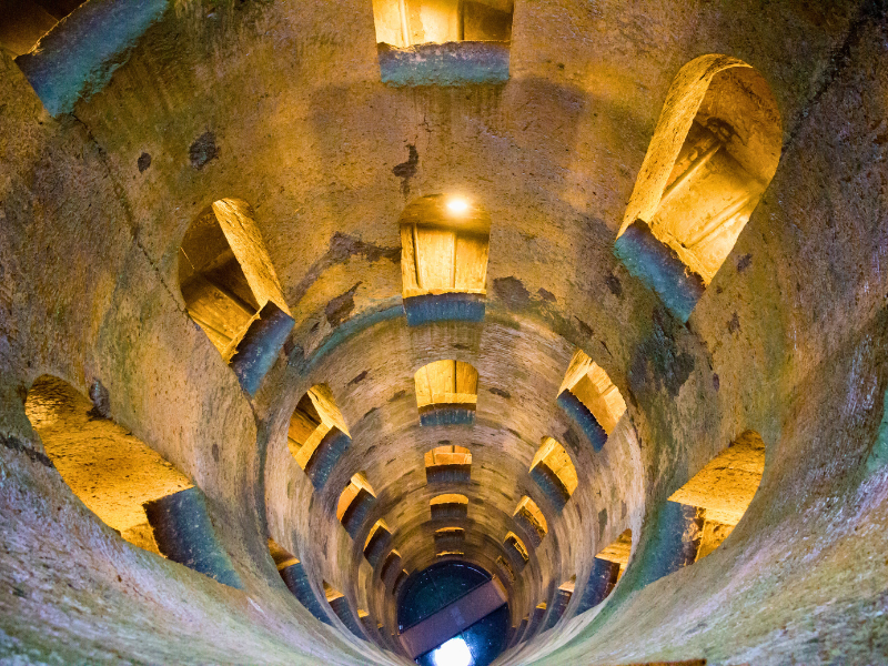 Looking down an ancient Italian well with levels and lit stone arches to bridge over water at bottom