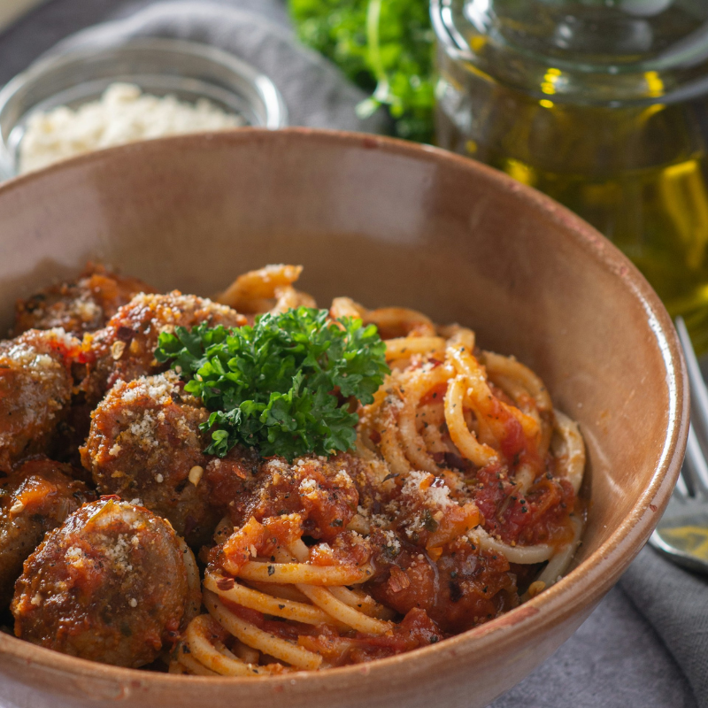 Bowl of spaghetti with tomato sauce, meatballs and parsley beside a jar of olive oil